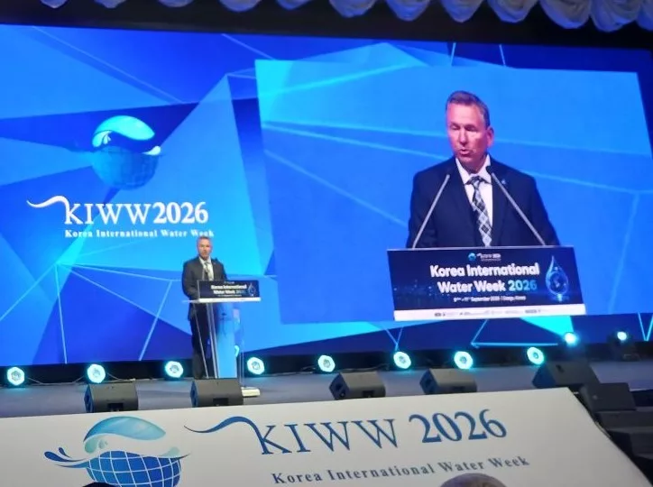 A man speaks at a podium on a stage with a videocast behind him.