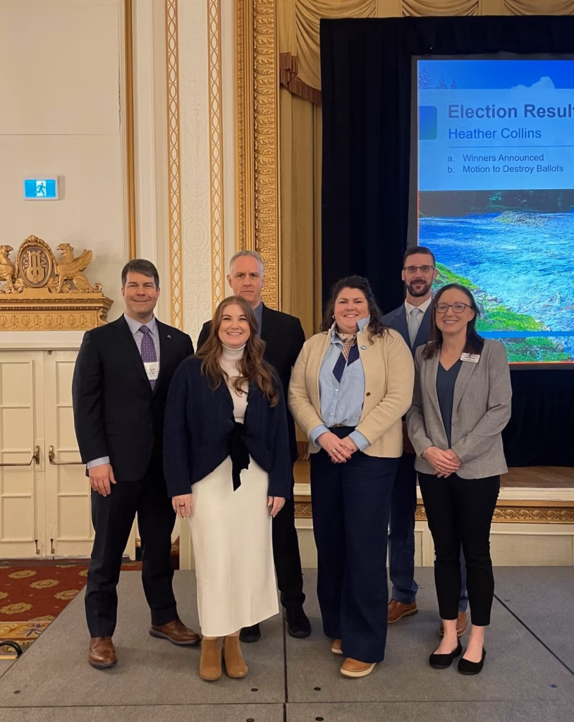 Six people stand for a posed shot in front of a presentation.